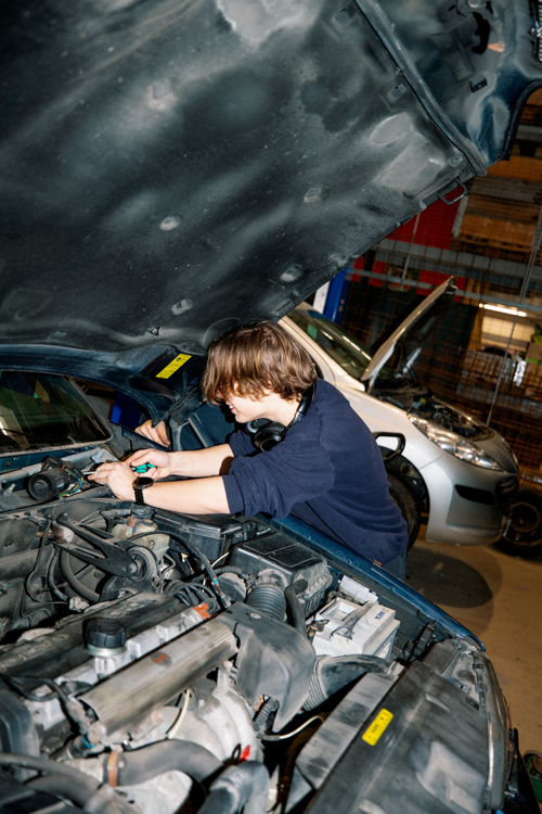 En studerende på GF1-uddannelsen på en erhvervsskole arbejder koncentreret under motorhjelmen på en bil i et værksted, Motorhjelm, Værksted, Bilmekanik, Automekaniker, Hands-on læring, Bilreparation,